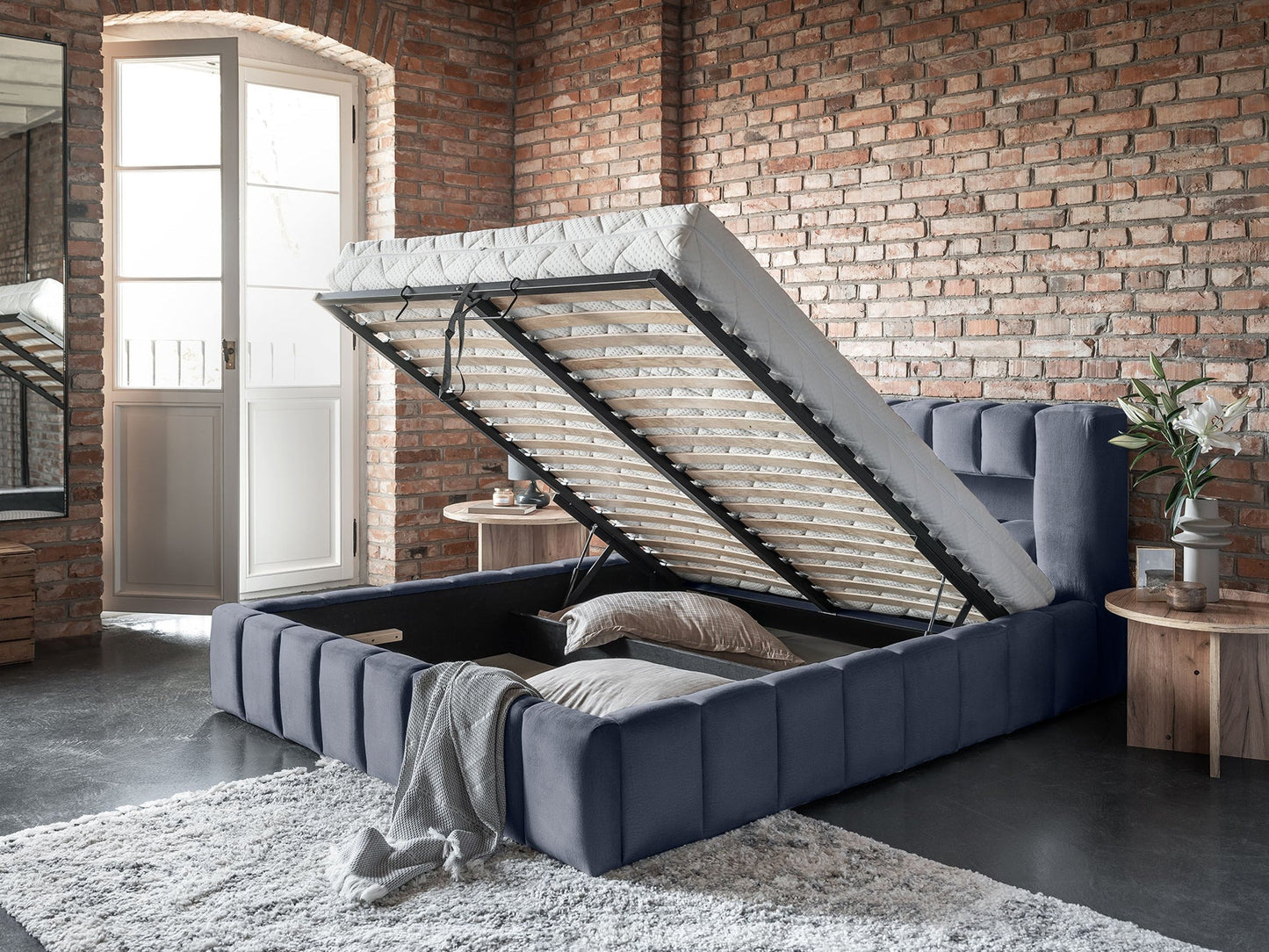 Łóżko Lamica 160x200 cm Monolith 97 ciemnoszary w lofcie z ceglaną ścianą, otwarty pojemnik na pościel.