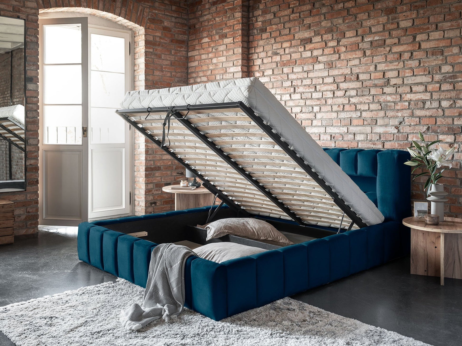 Granatowe łóżko Lamica 180x200 cm z otwartym pojemnikiem na pościel w industrialnej sypialni z ceglaną ścianą.