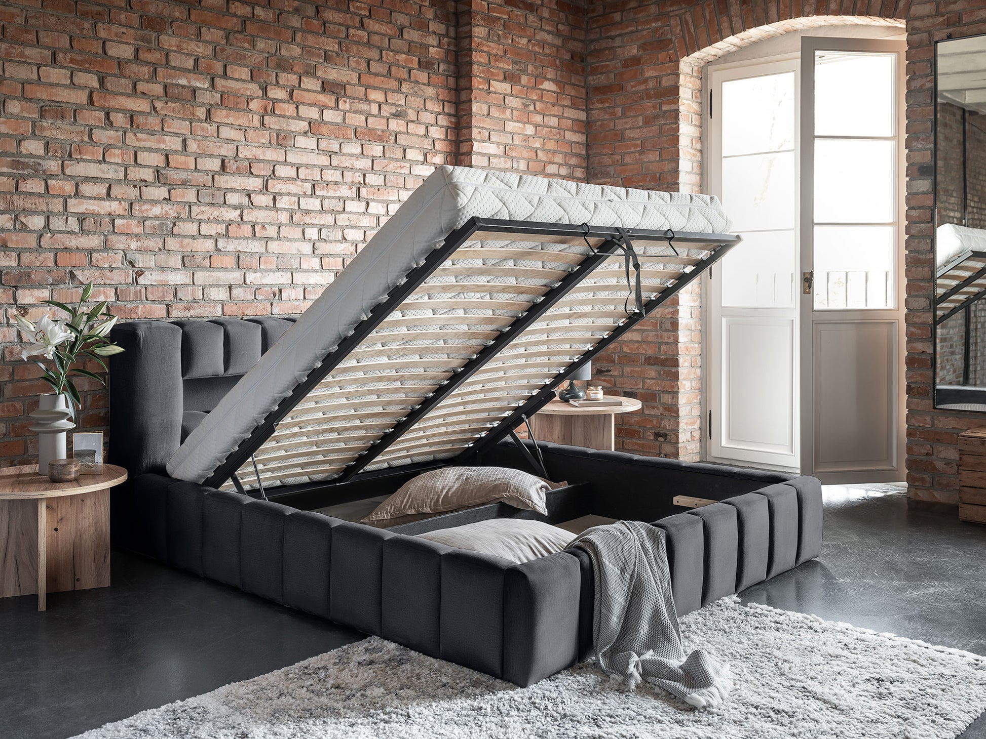 Łóżko tapicerowane Lamica 180x200 cm w kolorze Monolith 85 szary z pojemnikiem na pościel, pokazane w lofcie z ceglaną ścianą.