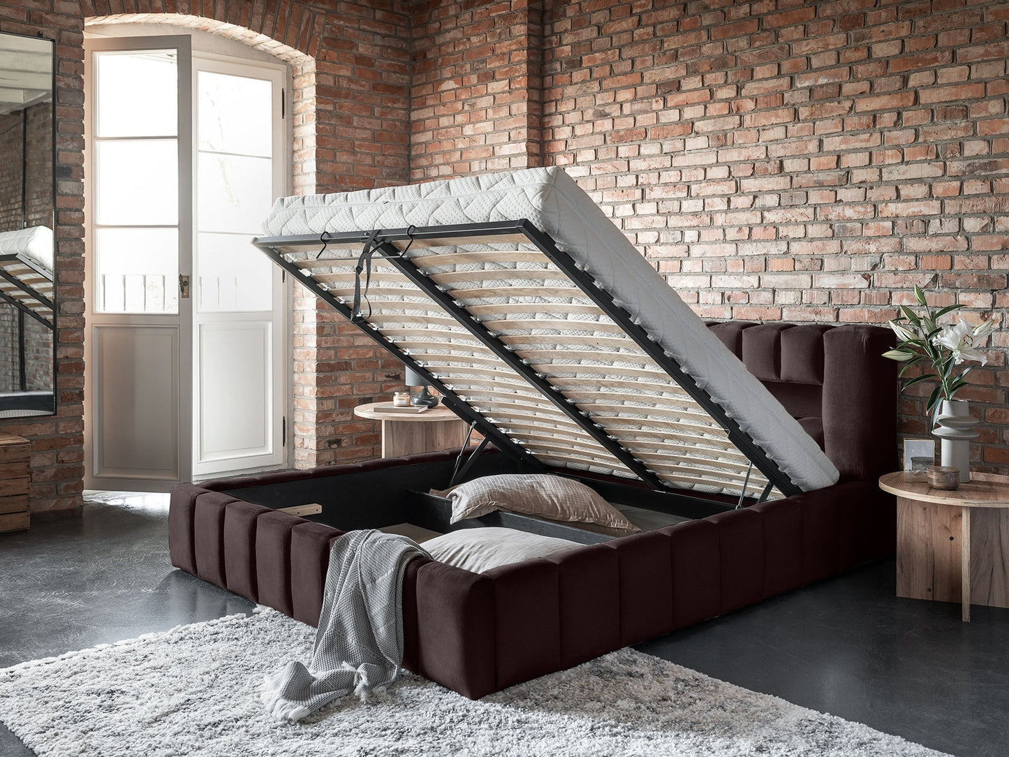 Perspektywa z boku na łóżko Lamica Monolith 29 brązowy (140 × 200 cm) z podniesionym stelażem i głębokim miejscem do przechowywania, na tle surowej, ceglanej ściany.