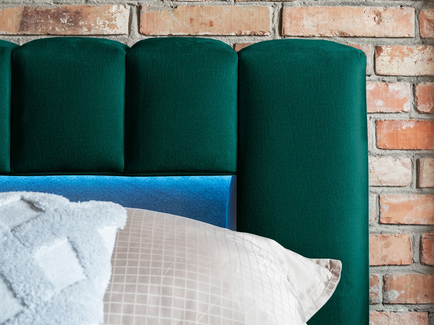 Zbliżenie na pikowany zagłówek łóżka Lamica II Monolith 37 butelkowozielony z wbudowanym oświetleniem LED na tle ceglanej ściany.