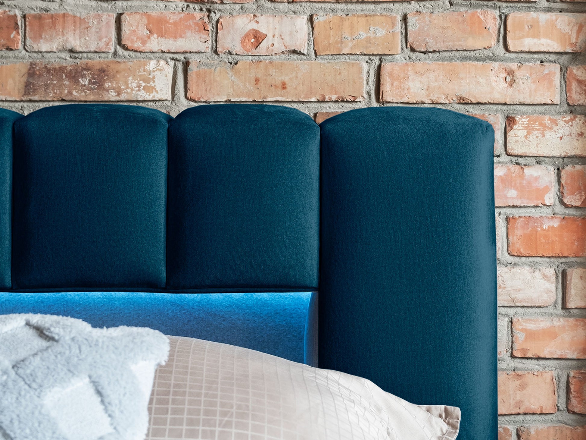 Zbliżenie na pikowany zagłówek łóżka Lamica II Monolith 77 granatowy z wbudowanym oświetleniem LED na tle ceglanej ściany.