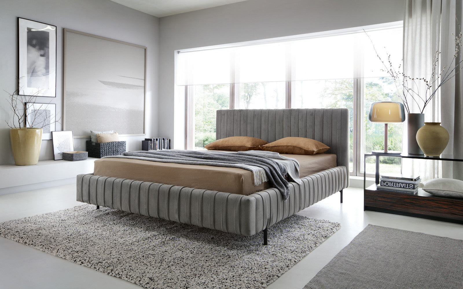 Łóżko Plissa 180×200 w kolorze Nube 05 ciemnoszarym w nowoczesnej, jasnej sypialni przy dużym oknie, z beżową pościelą i stolikiem nocnym z lampą.