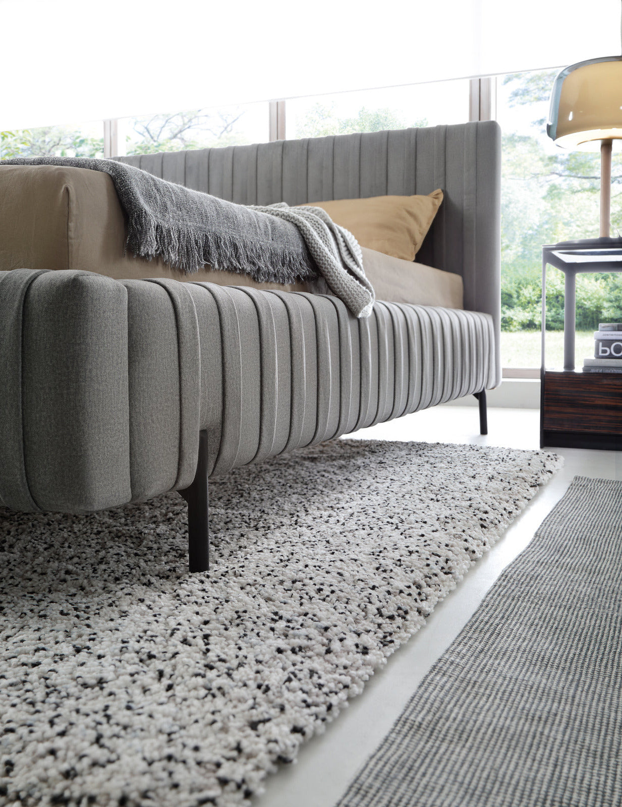 Widok boczny na dolną część ramy i smukłą, czarną nóżkę łóżka Plissa 180×200 w obiciu Nube 05 ciemnoszarym nad puszystym dywanem.