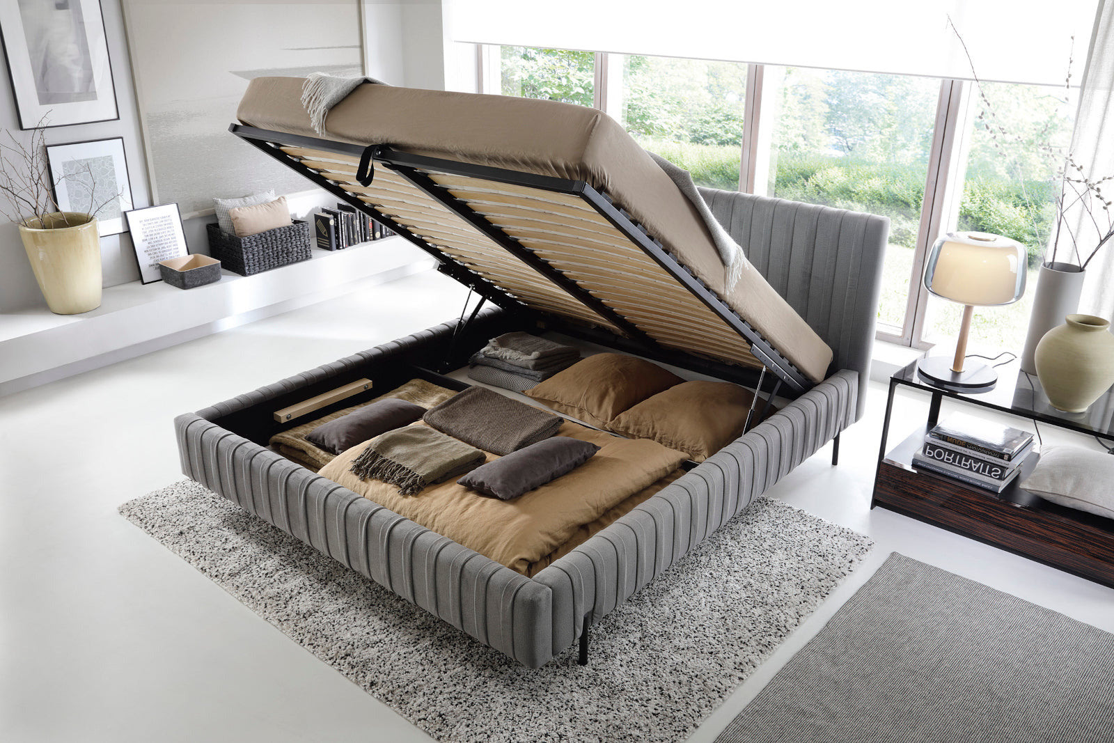 Łóżko Plissa 160×200 w obiciu Nube 05 ciemnoszarym ze stelażem uniesionym do góry, odsłaniającym pojemny schowek na pościel, ustawione na jasnoszarym dywanie.