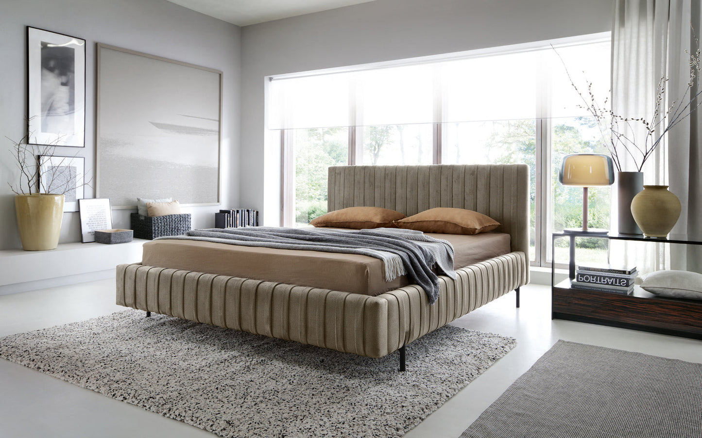 Łóżko Plissa 140×200 w kolorze Nube 20 beżowym w nowoczesnej, jasnej sypialni przy dużym oknie, z beżową pościelą, lampą i dekoracjami na stoliku nocnym.