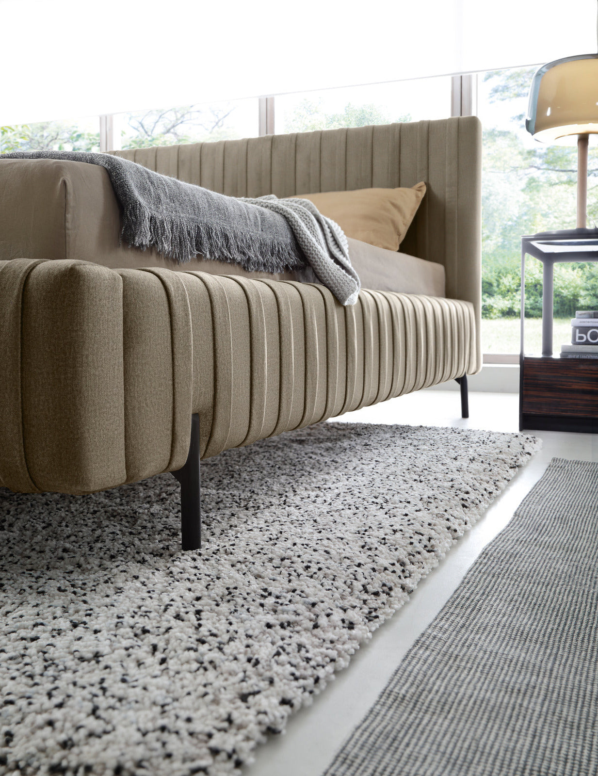 Widok boczny na ramę łóżka Plissa 160×200 w obiciu Nube 20 beżowym z cienką czarną nóżką, unoszącą je nad puszystym dywanem.