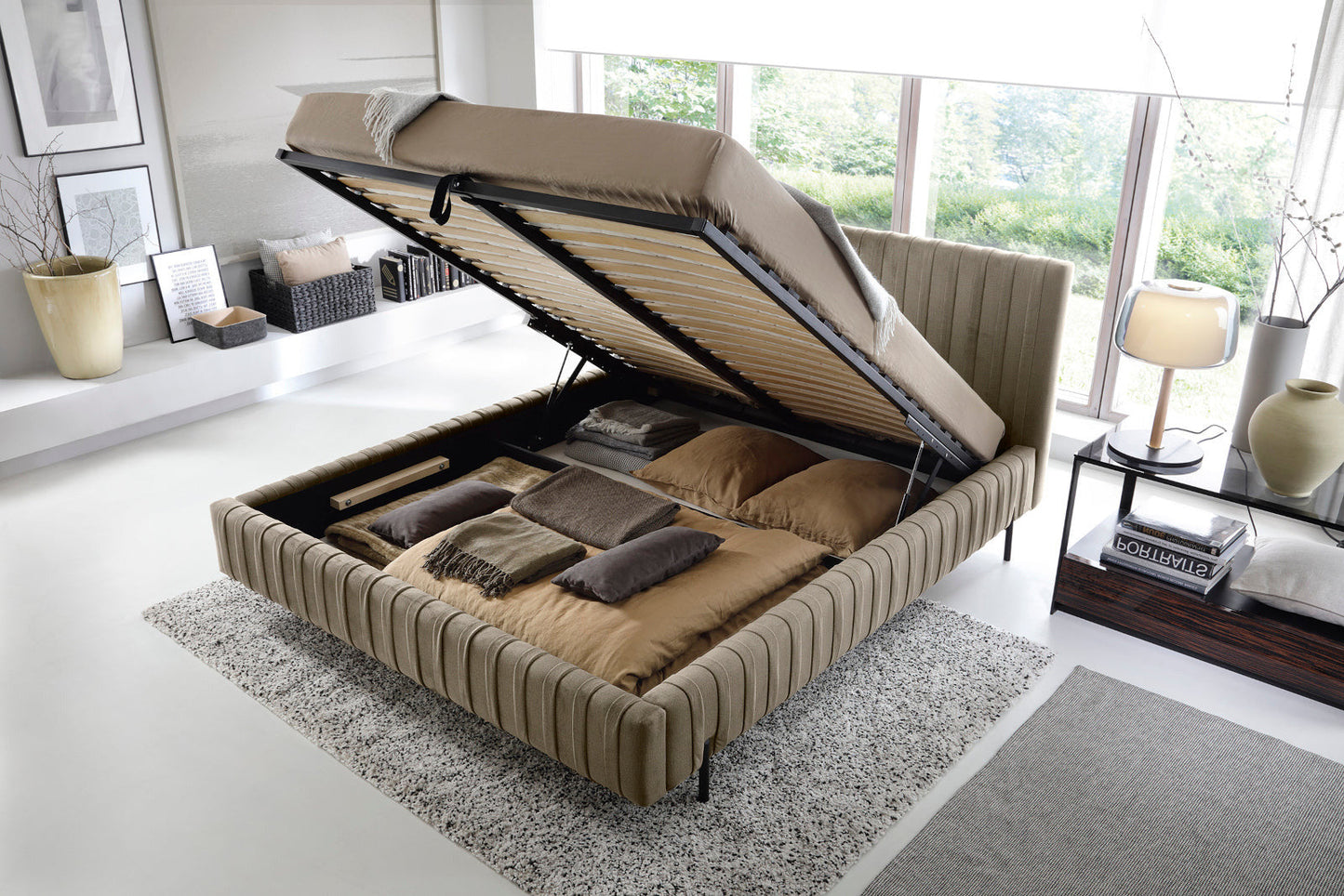 Łóżko Plissa 180×200 w obiciu Nube 20 beżowym ze stelażem uniesionym do góry, odsłaniającym pojemny schowek na pościel, ustawione nad dwoma dywanami.