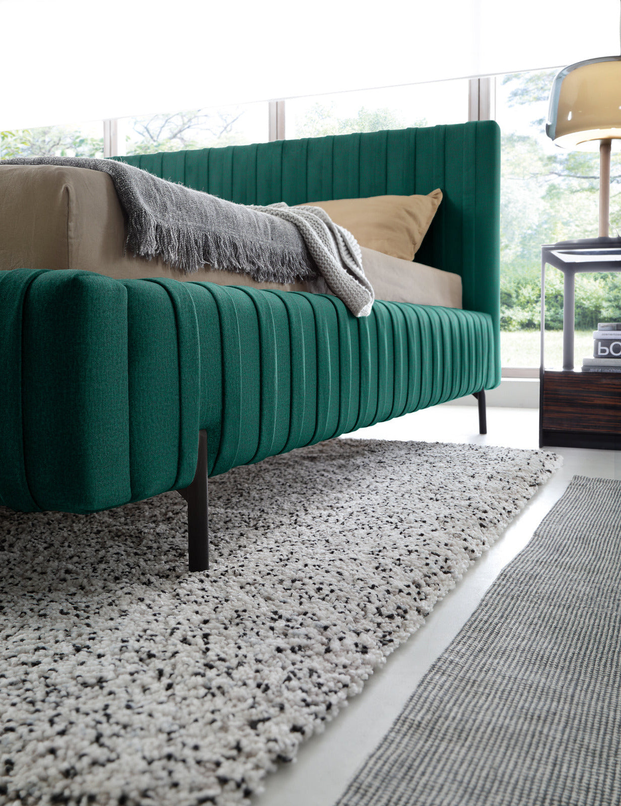 Widok boczny na dolną część ramy łóżka Plissa 140×200 w obiciu Nube 35 zielonym z cienką czarną nóżką nad puszystym dywanem.
