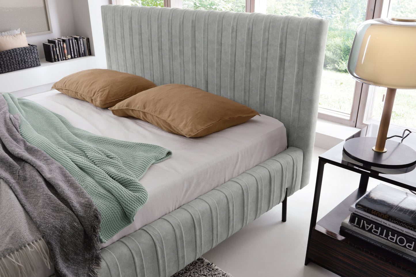 Zbliżenie na wezgłowie łóżka Plissa Relax 03 szary 180×200 w aranżacji wnętrza, z brązowymi poduszkami i dużą lampą na tle panoramicznego okna.