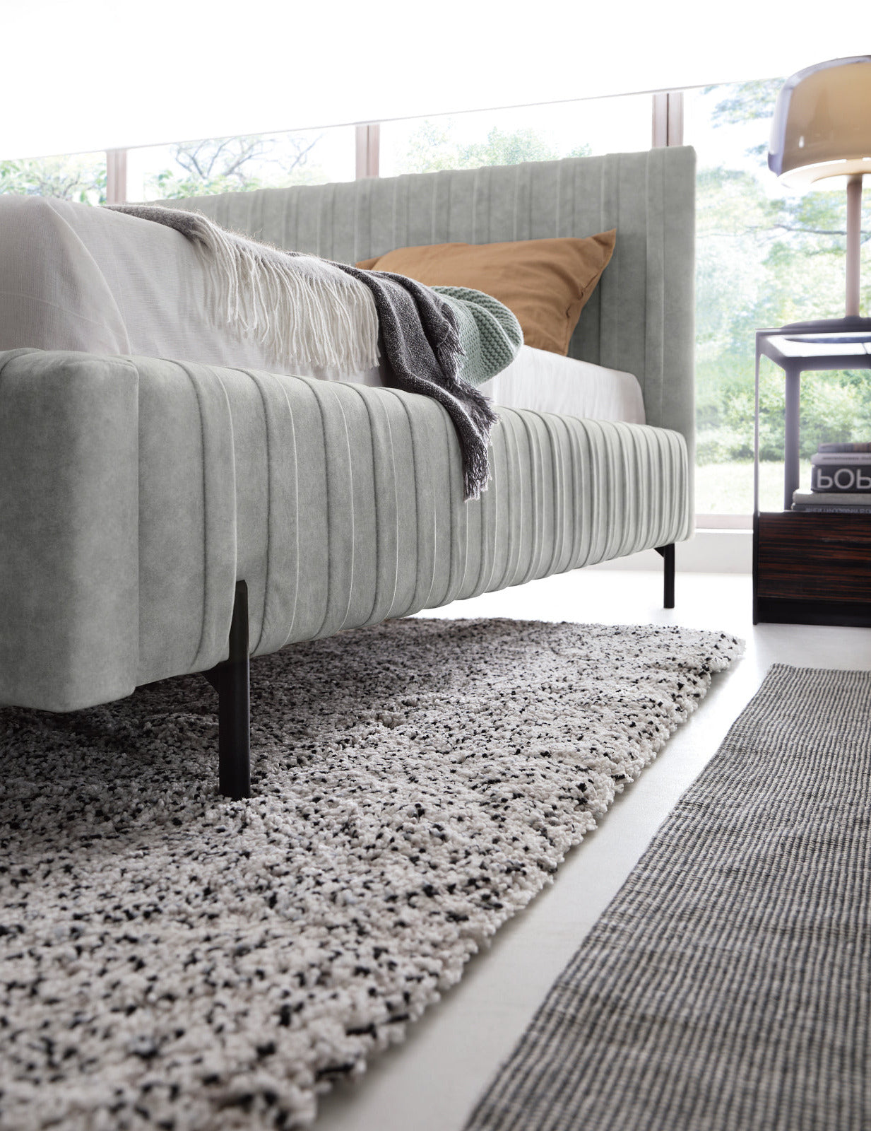 Boczna perspektywa łóżka Plissa 140×200 Relax 03 szary eksponująca profilowaną ramę i smukłe, czarne nogi na teksturowanym dywanie.
