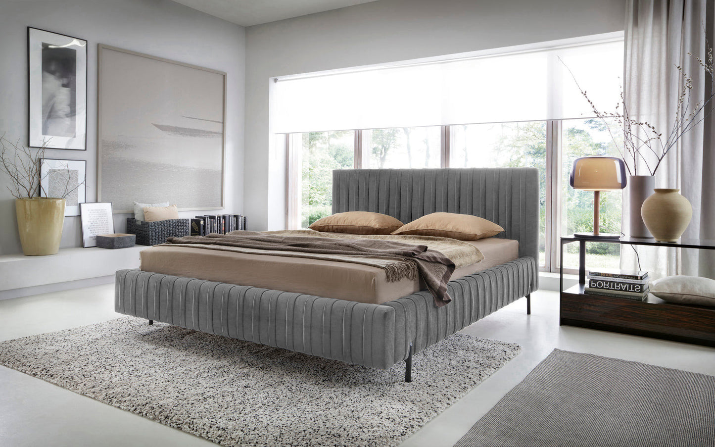 Łóżko Plissa 140×200 Velvetmat 04 szare w nowoczesnej sypialni przy dużym oknie, z beżową pościelą, brązowymi poduszkami i designerską lampą na szklanym stoliku nocnym.