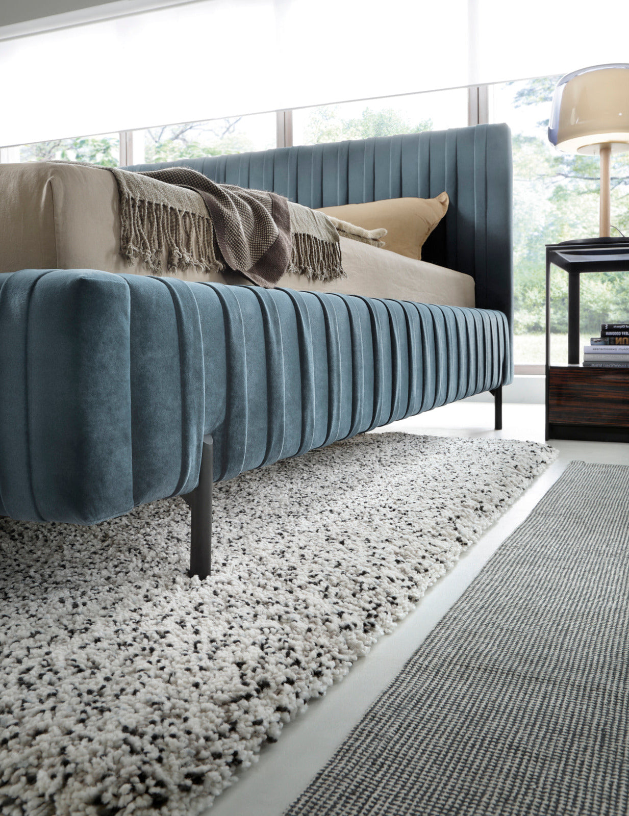 Widok boczny na dolną część ramy łóżka Plissa 180×200 w obiciu Velvetmat 100 jasnoniebieskim z cienką czarną nóżką, nad warstwą puszystego dywanu.