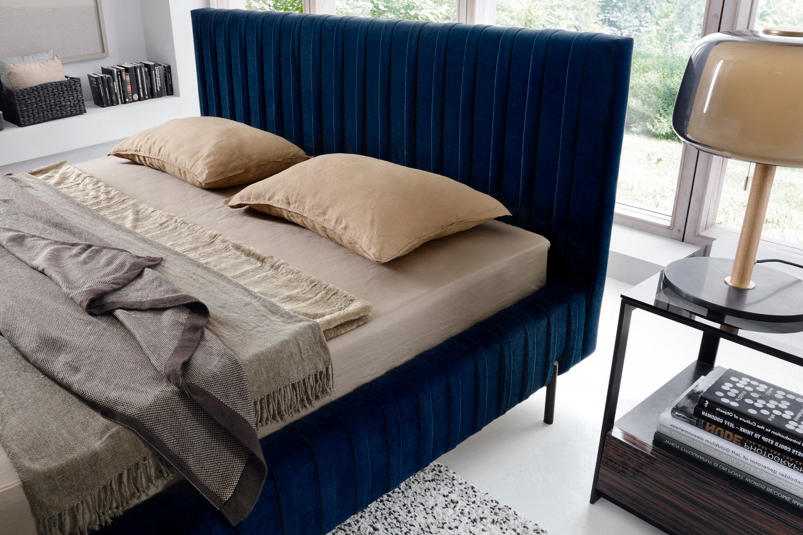 Zbliżenie na wysokie, pionowo karbowane wezgłowie łóżka Plissa 140×200 Velvetmat 40 granatowego z beżowymi poduszkami i lampą na tle dużego okna.