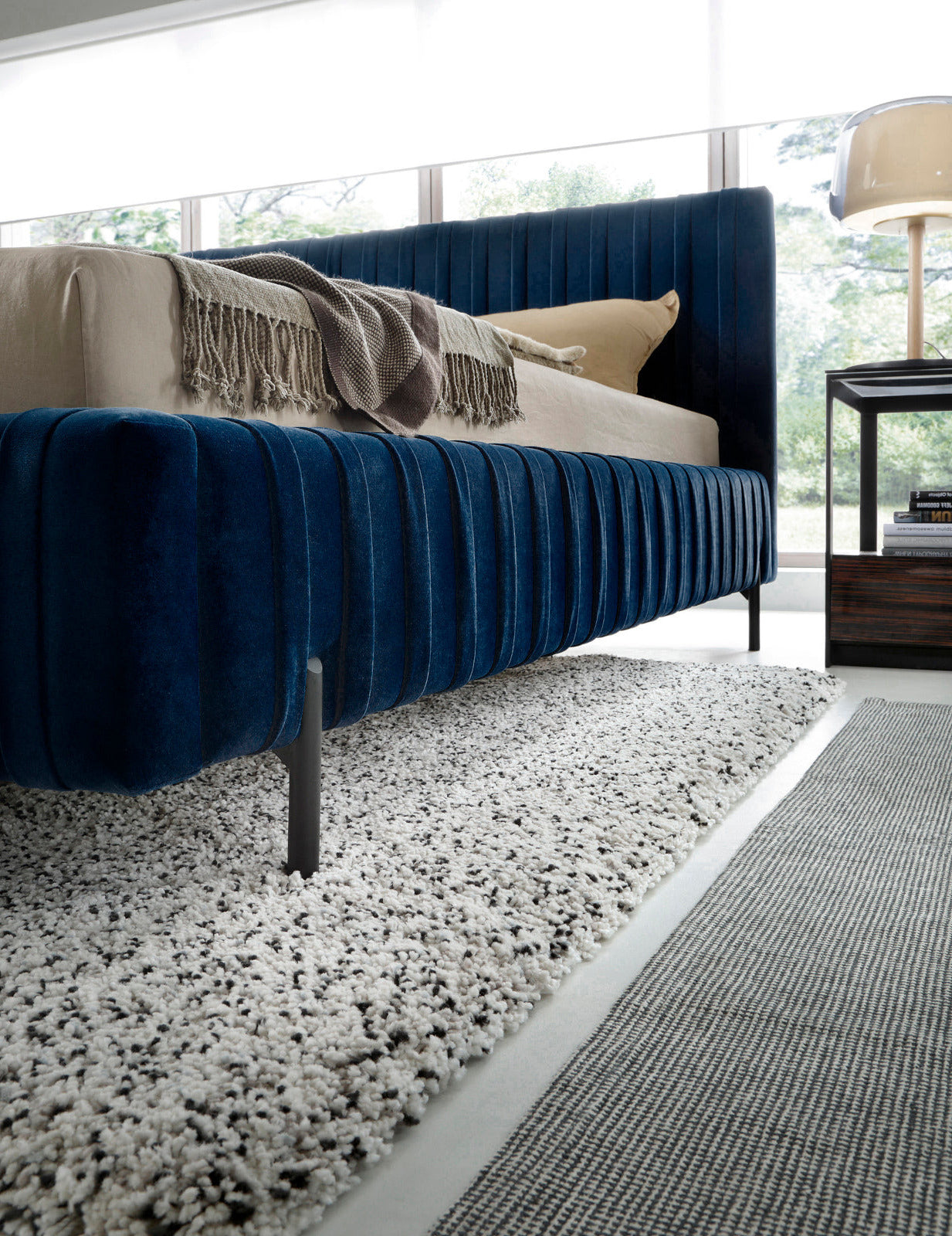 Boczna perspektywa łóżka Plissa 180×200 Velvetmat 40 granatowy na teksturowanym dywanie, eksponująca profilowaną ramę i smukłe, czarne nogi.