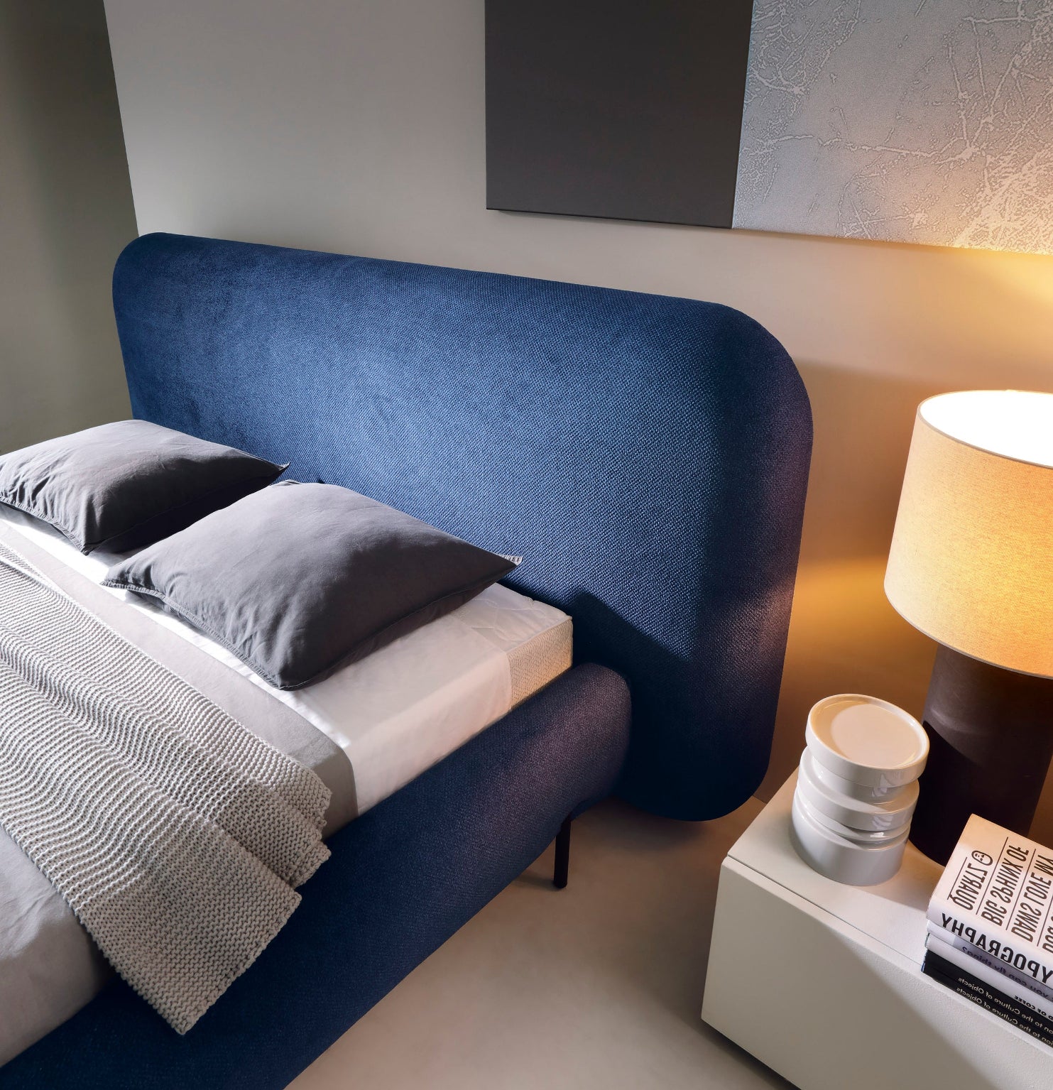 Zbliżenie na zaokrąglony zagłówek niebieskiego łóżka tapicerowanego Paoli 160×200 cm z poduszkami i lampą nocną