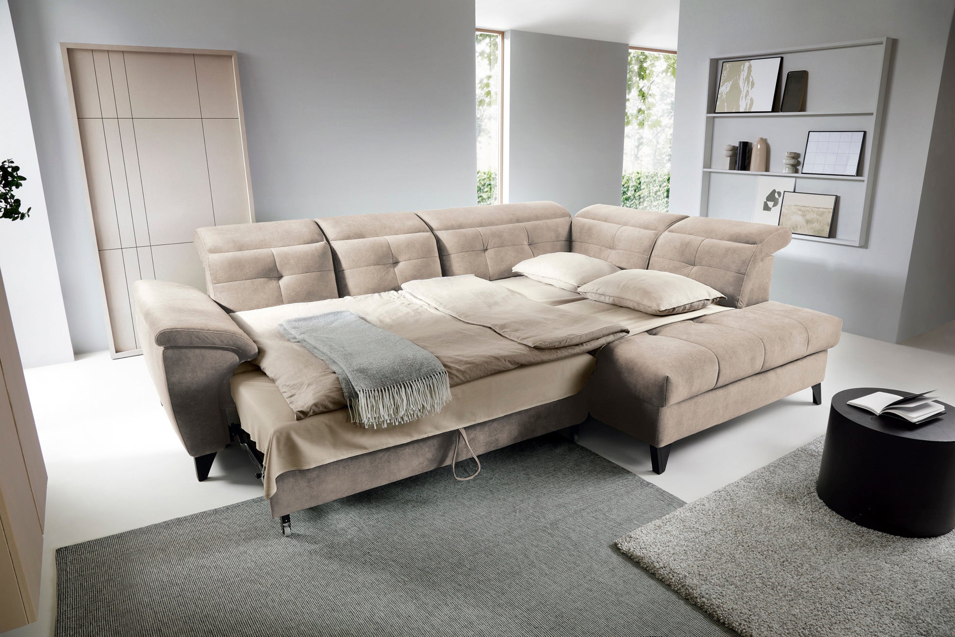 Narożnik Inferne Relax 20 beżowy w układzie prawym rozłożony jako łóżko, z różnie ustawionymi zagłówkami i pościelą na siedzisku, w jasnym wnętrzu z dywanami i obrazami na ścianie.