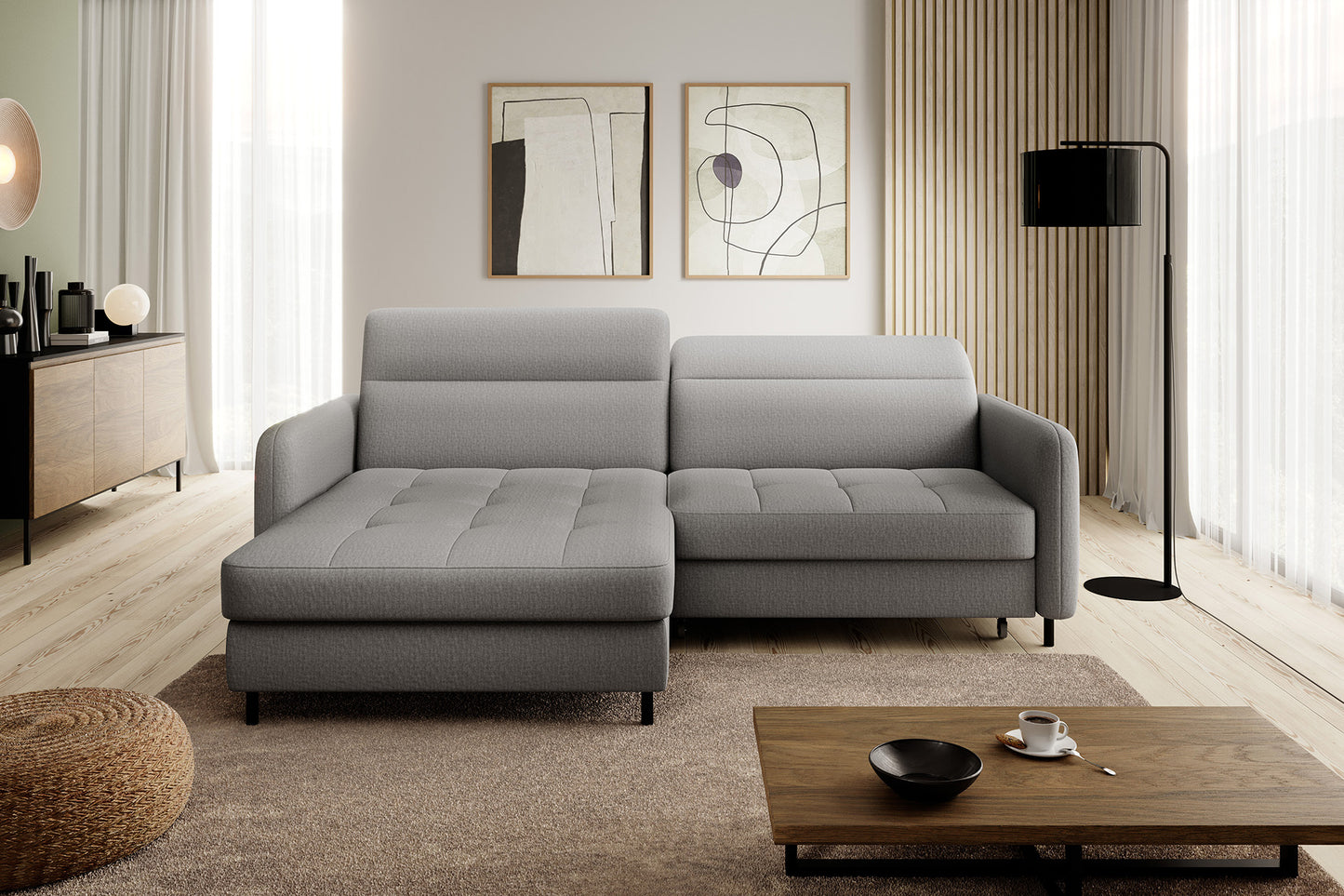 Nowoczesny salon z narożnikiem Gomsi Manhattan 03 jasnoszary w układzie lewym, umieszczonym obok czarnej lampy podłogowej i drewnianej komody przy dużym oknie.