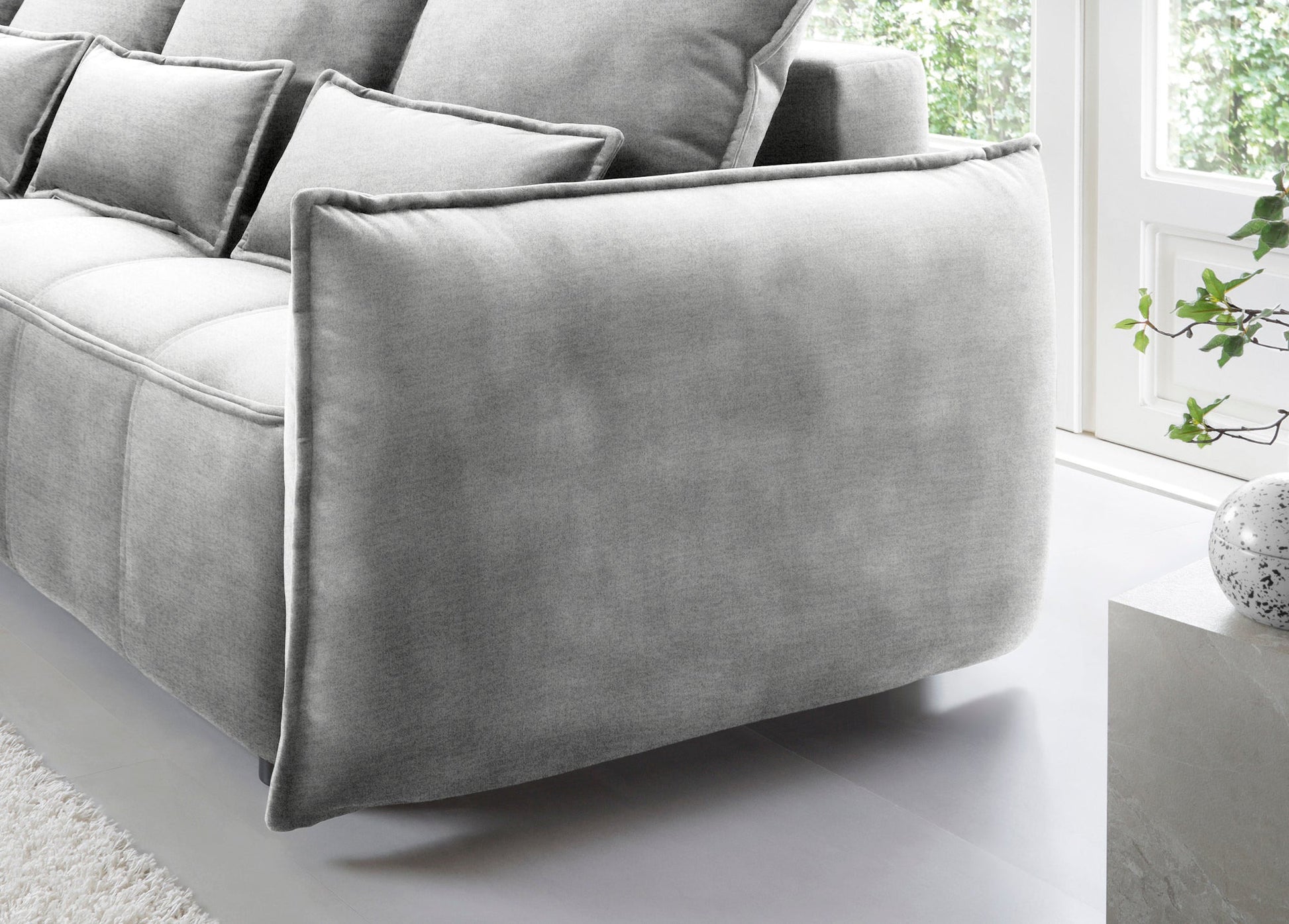 Detal zaokrąglonego podłokietnika i tylnej części narożnika Campo w układzie lewym, tapicerka Nube 03 jasnoszara na tle okna.