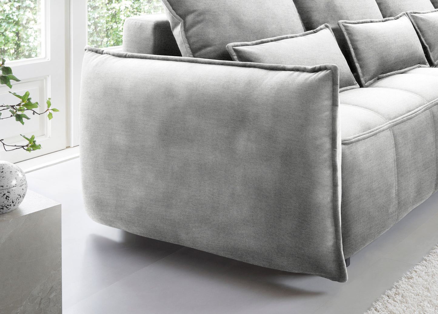 Detal zaokrąglonego podłokietnika i tylnej części narożnika Campo w układzie prawym, tapicerka Nube 03 jasnoszara na tle okna.