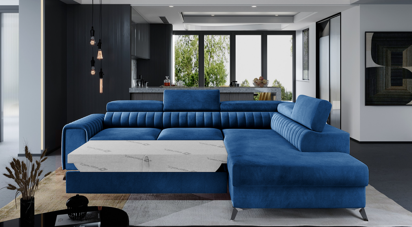 Niebieski narożnik Laurence Kronos 09 w nowoczesnym salonie z dużymi przeszkleniami i elegancką kuchnią w tle. Mebel posiada funkcję spania oraz regulowane zagłówki, zapewniając komfort i funkcjonalność w codziennym użytkowaniu.