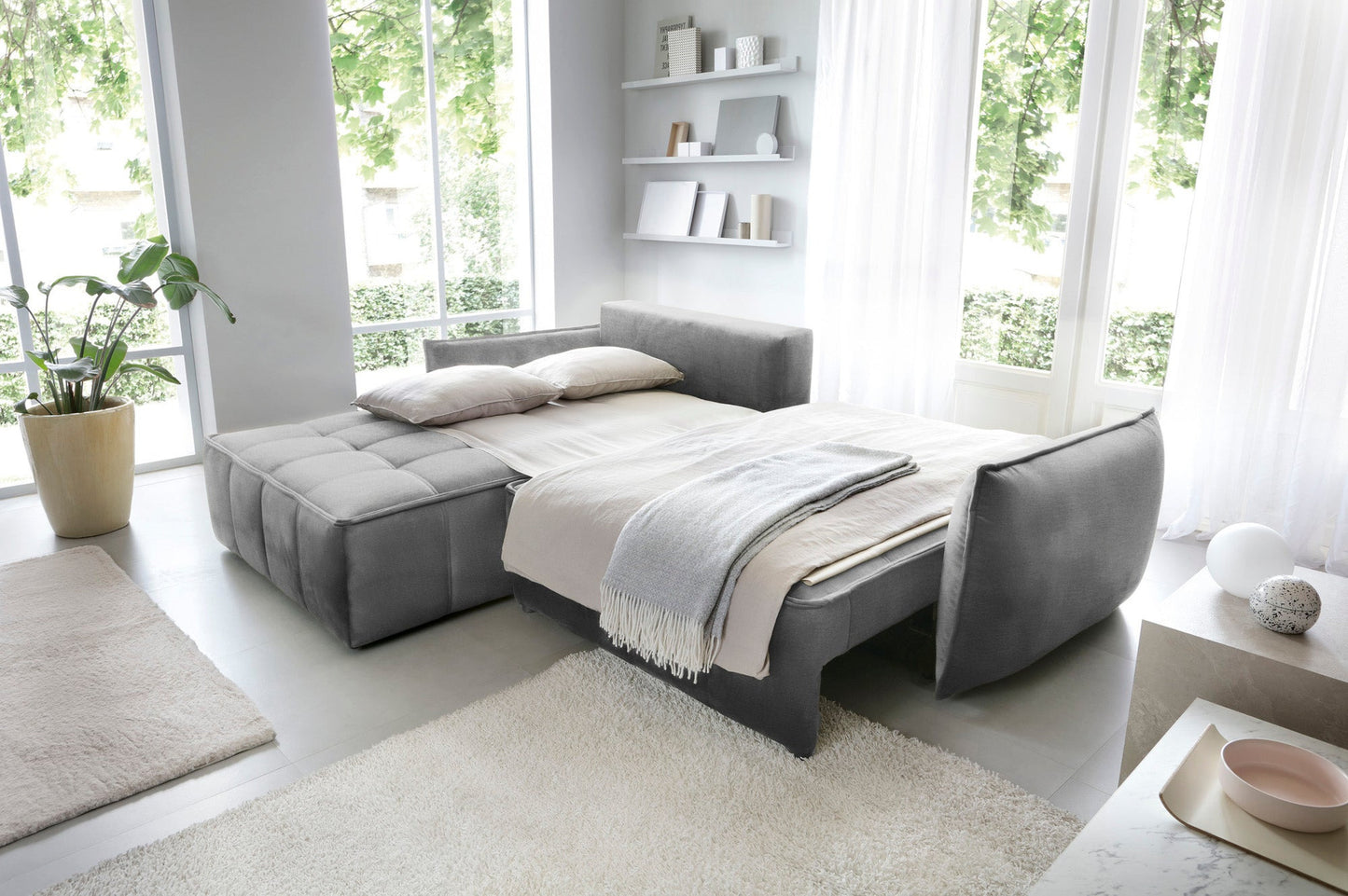 Narożnik Campo w układzie lewym, tkanina Nube 04 szary, rozłożony jako łóżko z narzutą i poduszkami w przestronnym, jasnym pomieszczeniu.