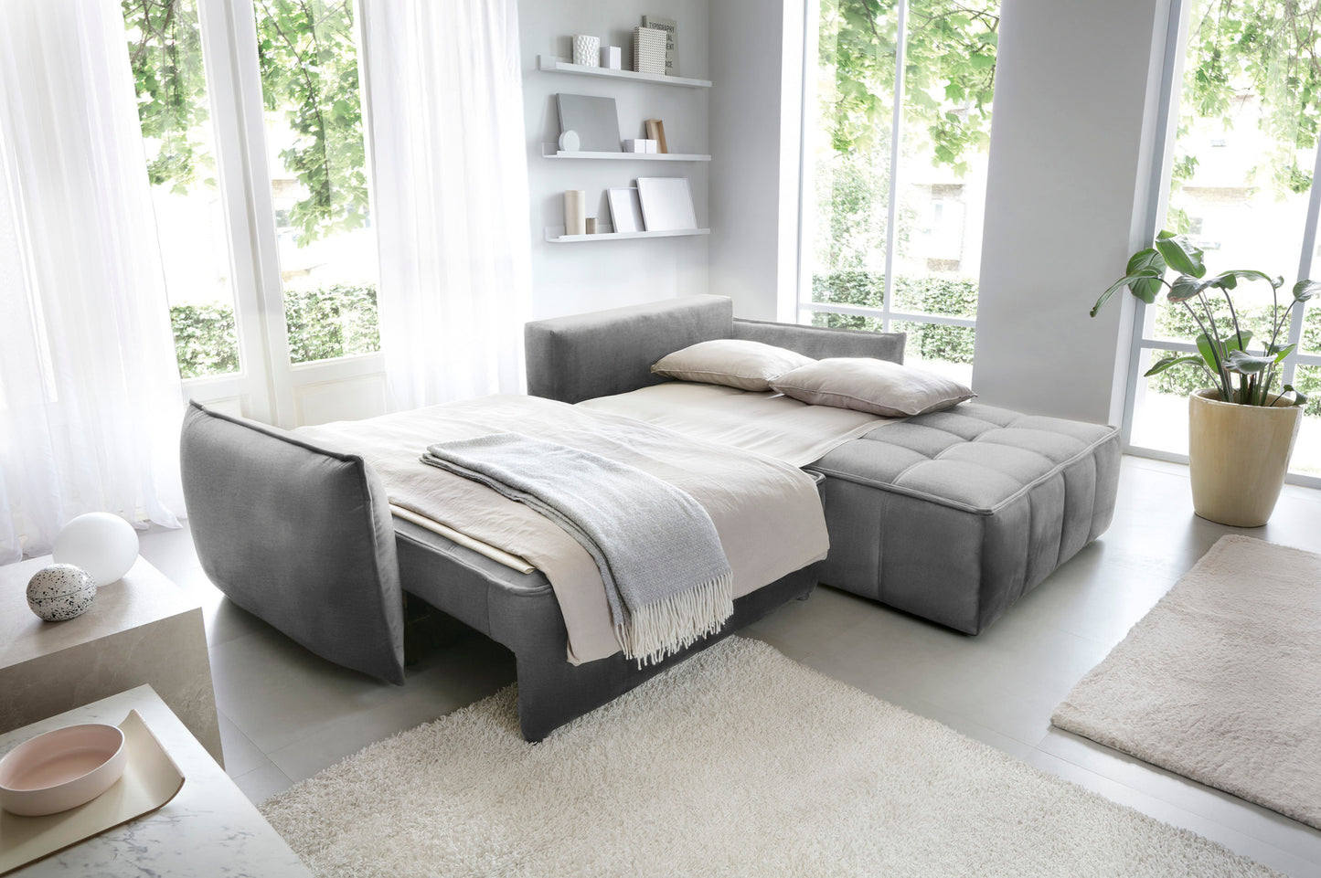 Narożnik Campo w układzie prawym, tkanina Nube 04 szary, rozłożony jako wygodne łóżko z narzutą i poduszkami w przestronnym, jasnym pomieszczeniu.