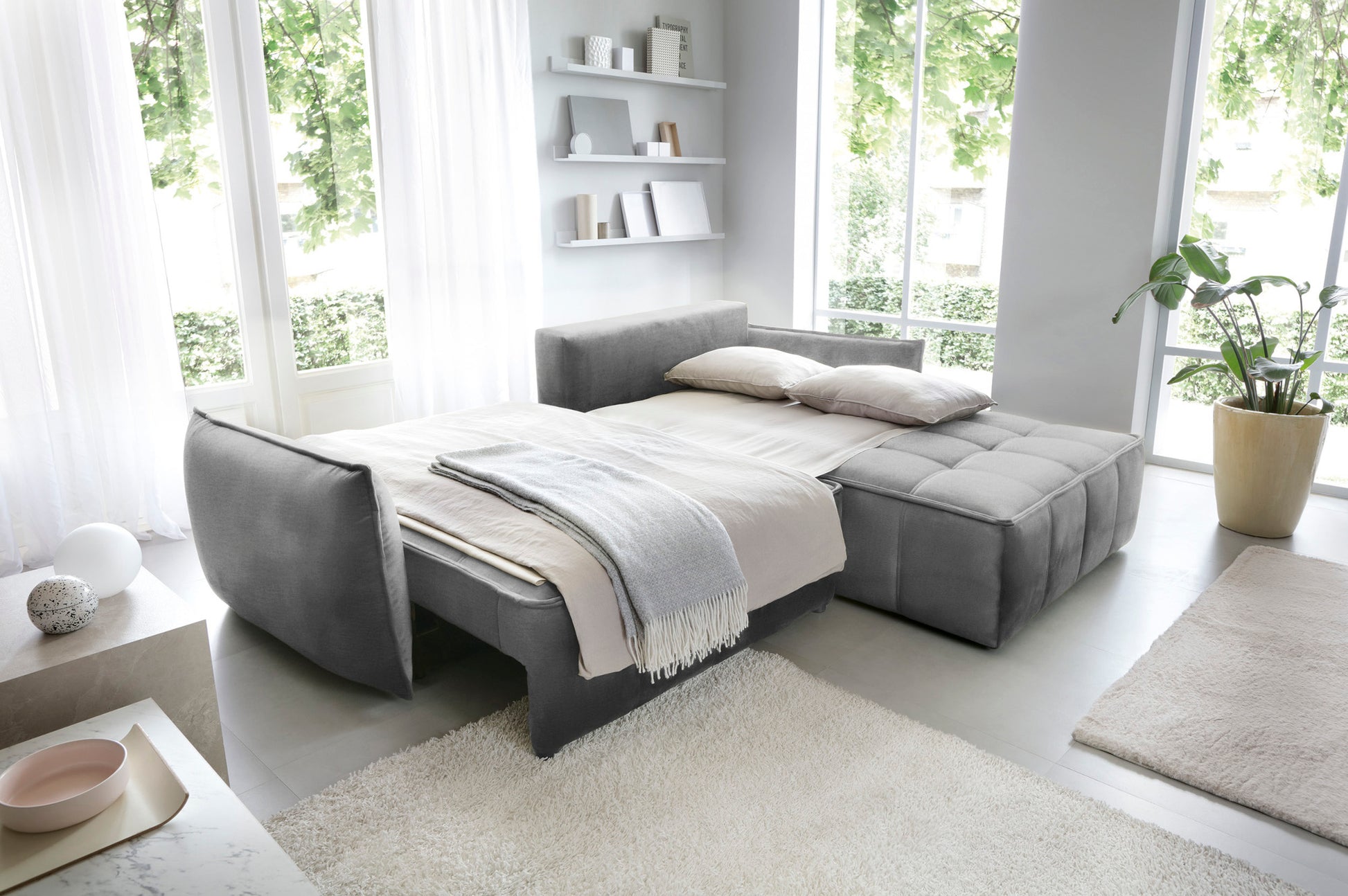 Narożnik Campo w układzie prawym, tkanina Nube 04 szary, rozłożony jako wygodne łóżko z narzutą i poduszkami w przestronnym, jasnym pomieszczeniu.
