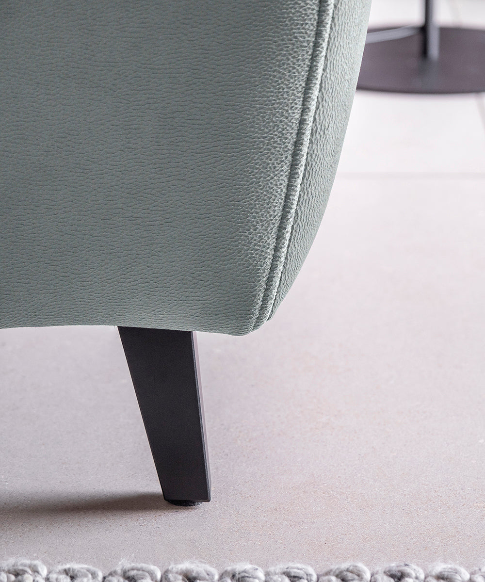 Fragment z nóżką narożnika Noble w jasnoszarej tkaninie Savoi 100 z dekoracyjnymi przeszyciami i subtelną strukturą materiału. Elegancki narożnik do salonu inspirowanego stylem contemporary, soft loft i japandi.