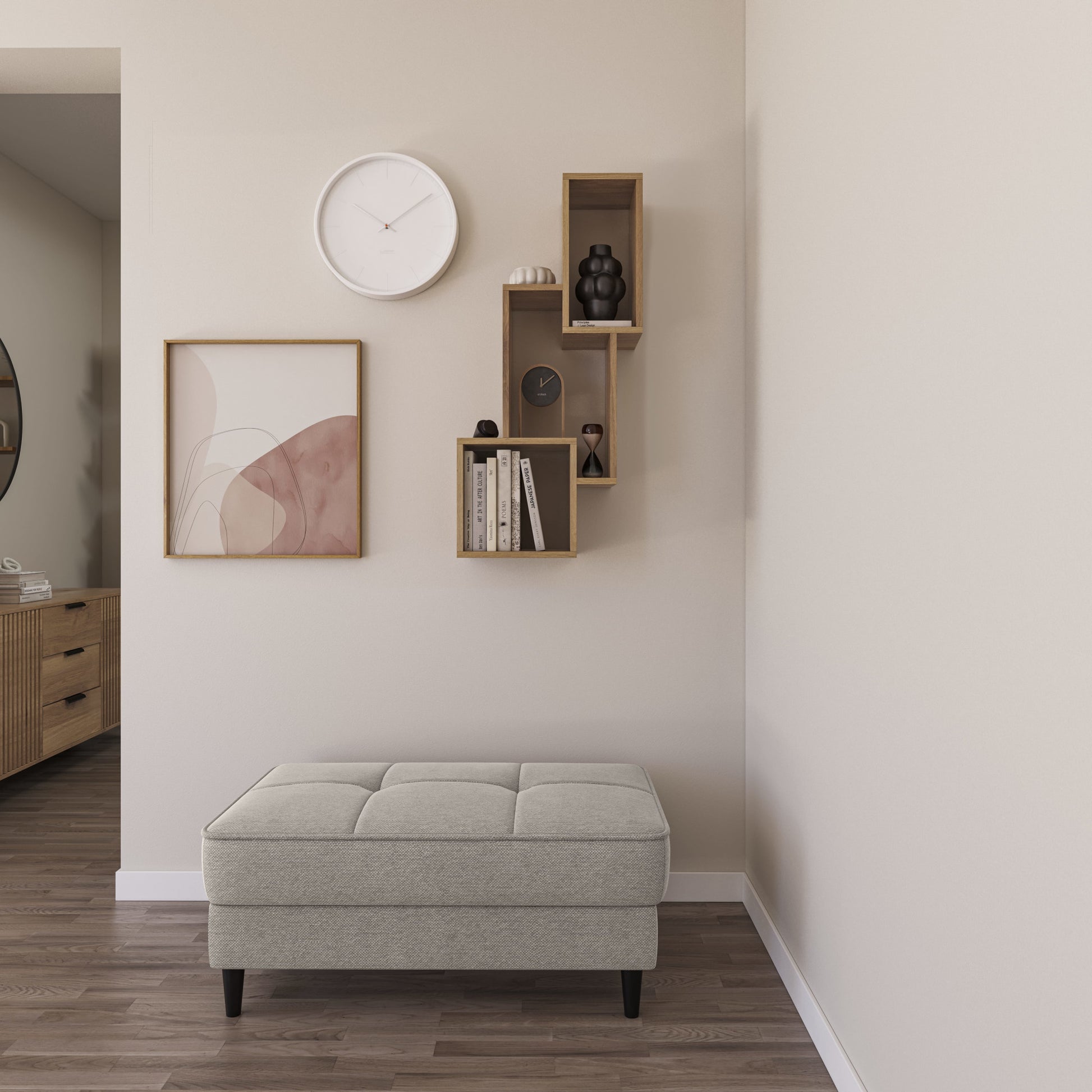 Półka Alitty dąb artisan zawieszona na jasnej ścianie nad pikowanym pufem; na półkach książki, zegar i dekoracyjne figurki wstępnie udekorowane abstrakcyjną grafiką oraz białą lampą.