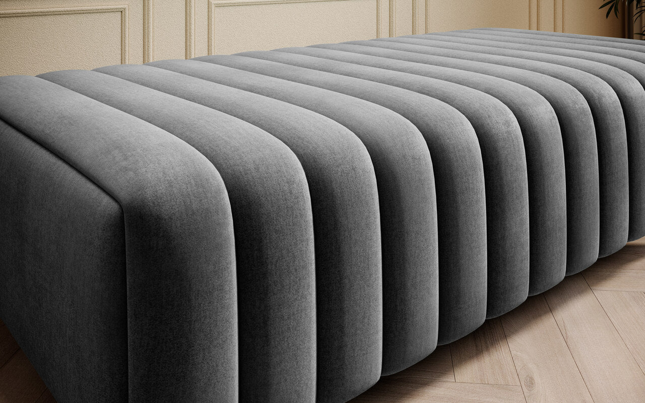 Elegancka pufa do sypialni Laurence Nube 05 ciemnoszara ustawiona przed łóżkiem jako wygodne miejsce do siedzenia, odkładania tekstyliów i uzupełnienia nowoczesnej aranżacji.