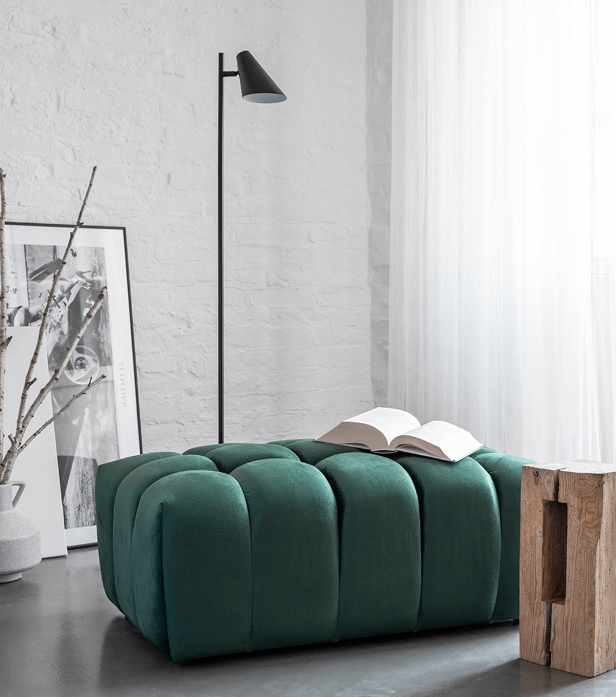 Pufa Lazaro w głębokim zielonym kolorze Kronos 19 ustawiona przy białej ceglanej ścianie i oknie z lekką zasłoną. Na siedzisku leży otwarta książka, a obok znajdują się minimalistyczna lampa podłogowa i stolik z drewna.