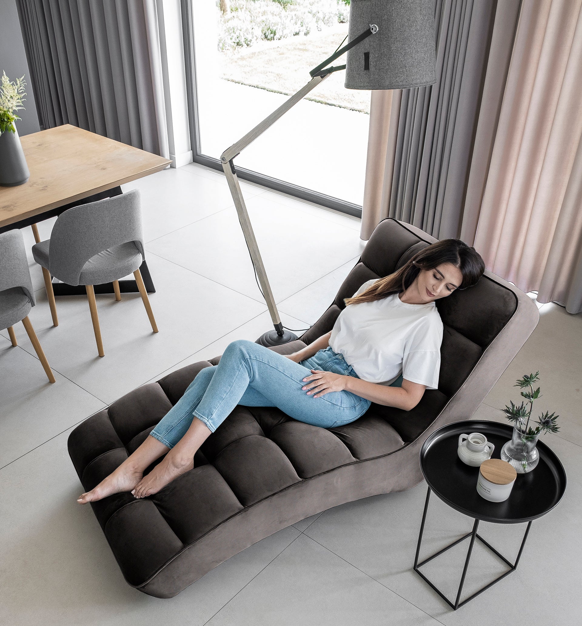 Kobieta w białej bluzce i dżinsach odpoczywa na ciemnoszarym szezlongu Loren w eleganckim salonie z dużym oknem.