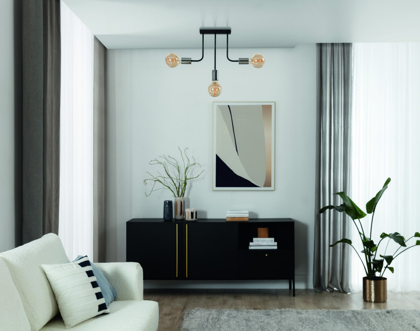 Żyrandol Indigeo 48x48 cm z trzema źródłami światła w nowoczesnym salonie z jasnymi zasłonami i czarną komodą.
