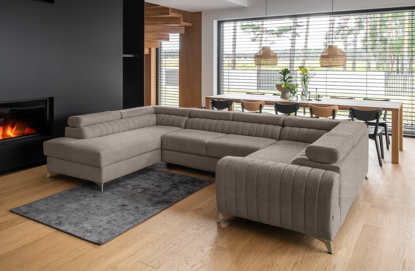 Przestronny salon z narożnikiem Louis w kolorze Poco 07 beżowy w układzie lewym, ustawionym przy kominku na drewnianej podłodze, z dużym oknem i jadalnią w tle.